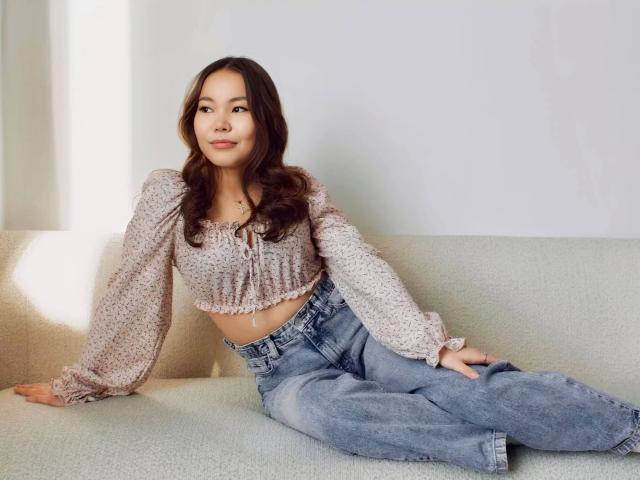 Sakura-hot has medium tits, long brown hair, wearing a floral crop top and high-waisted blue jeans, sitting on a light green couch, looking at the camera.