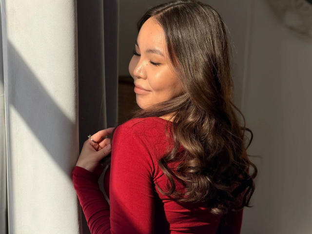 Sakura-hot with long, wavy brown hair in a red long-sleeve top, standing by a sunlit window, eyes closed, smiling softly, sunlight highlighting her face and chest.