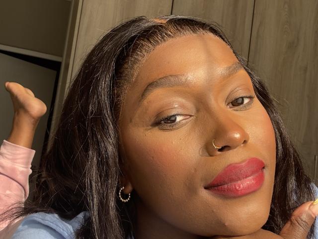 LaToyah ha la pelle marrone scuro, rossetto rosso, un piercing al naso e lunghi capelli neri con una piccola treccia sulla parte superiore. Il suo piede è sollevato, mostrando una posa rilassata..