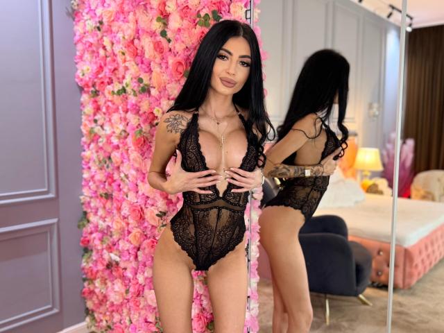 MadisonDesire with long black hair, wearing a black lace bodysuit, covering her tits with her hands, standing in front of a mirror with pink floral wall.