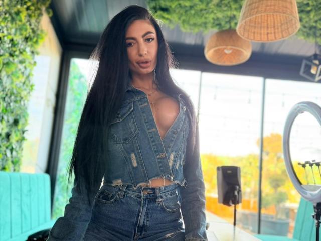 MadisonDesire with long black hair, wearing a blue denim crop top and high-waisted jeans, showing her small tits, standing in a bright room with a ring light.