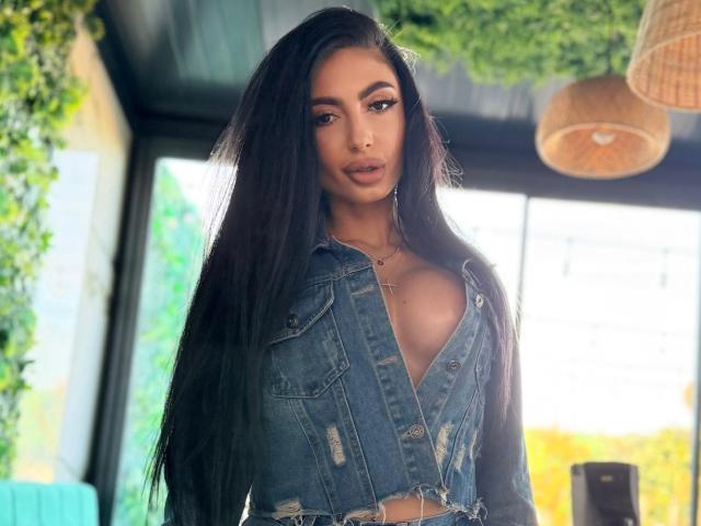 MadisonDesire with long black hair, wearing a blue denim crop jacket, showing her tits, and a cross necklace, standing in a sunlit room with greenery.