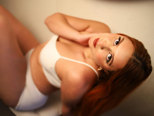 Antonela Salazar, con cabello rojo, vestida con un sostén y bombacha blancos, mirando hacia arriba con una mirada seductora, su mano cerca de su boca, iluminación suave, ligeramente borroso, sentada sobre una superficie..