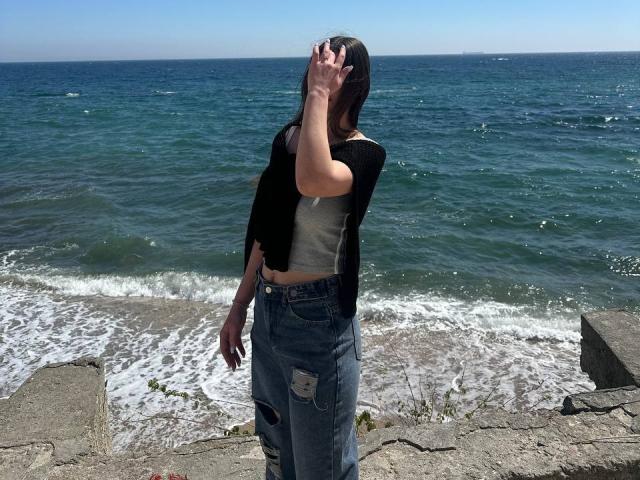 Omella's standing by the ocean, wearing a black cardigan, white crop top, and ripped blue jeans, with her hand covering her head, showing off her tits and ass.