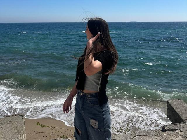 Omella with long brown hair, black jacket, gray top, and ripped jeans stands by the ocean, hand in hair, looking at the water. Her ass is slightly visible.