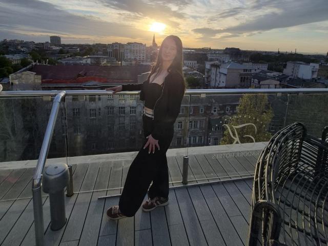 Omella with big tits, black hoodie, and black pants, standing on a rooftop at sunset, wearing sandals, smiling, hands on hips, metal railing and chair on wooden deck.