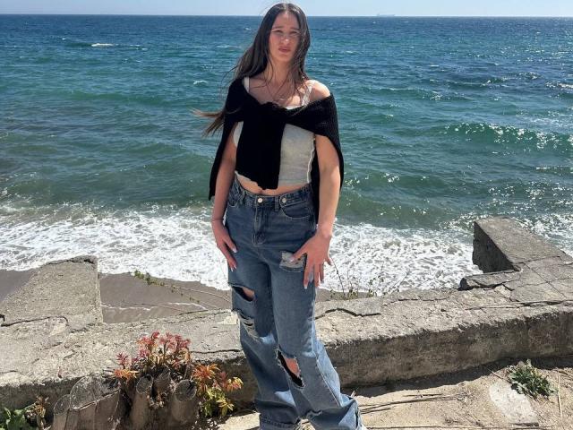 Omella stands by the ocean, wearing a black and white off-shoulder top and ripped blue jeans, with long brown hair blowing in the wind. Her tits are small.