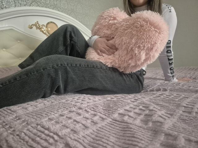 ZoieHurt sentada em uma cama com um travesseiro rosa fofo em formato de coração, vestindo uma camisa branca de manga comprida e calças cinzas escuras, com o bumbum ligeiramente levantado..