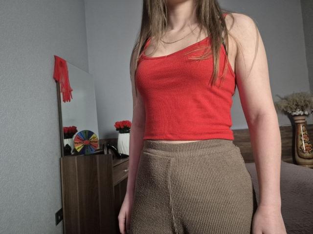 ZoieHurt with long brown hair, red tank top, and brown ribbed skirt, standing in a room, showing her tits and ass. Her face is out of frame.