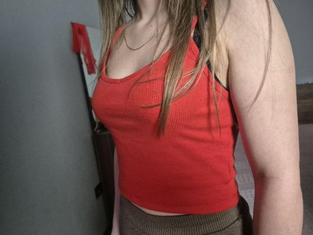 ZoieHurt with blonde hair, wearing a red ribbed tank top that shows her tits, and brown knit pants. She has a gold necklace and her face is out of frame.