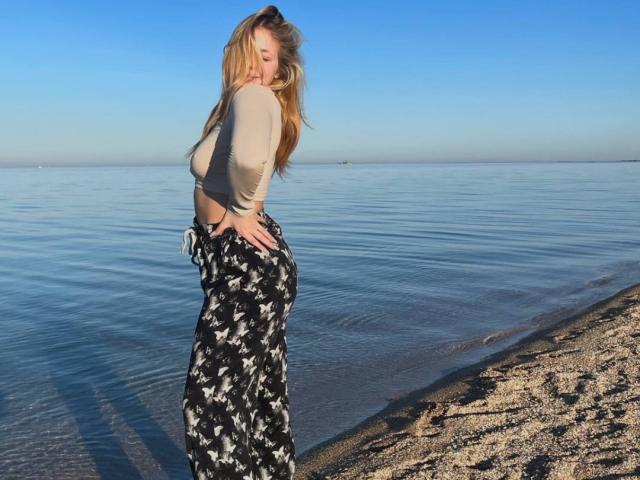 AnyaHoehn with blonde hair, small tits, wearing a beige top and black floral pants, standing on a pebble beach, facing left, hands on hips, water and clear sky.