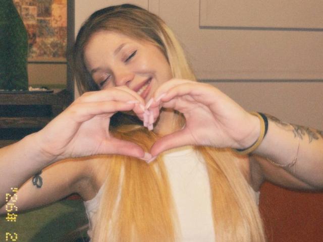 AnyaHoehn with long blonde hair, smiling, making a heart shape with her hands, showing her pink nipple, wearing a white sleeveless top, and a green bracelet.