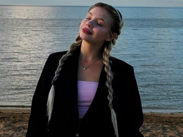 AnyaHoehn with braided blonde hair, eyes closed, smiling, wearing a black blazer over a pink top, standing by a calm blue sea, looking relaxed and happy.