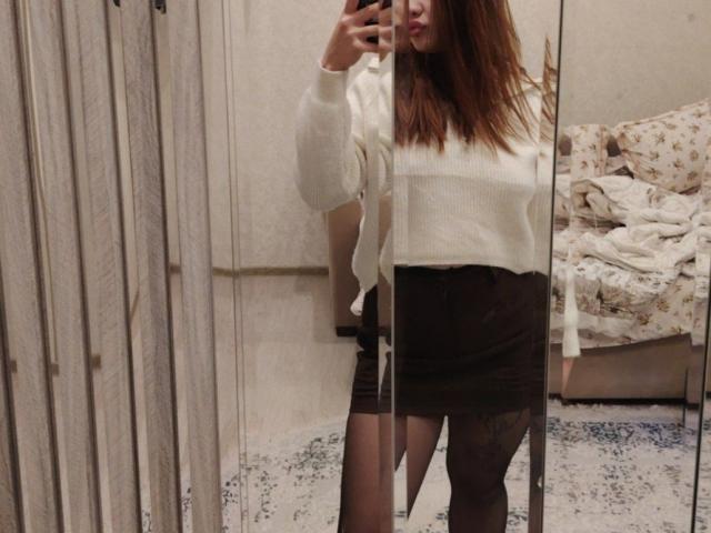 HotVibeK with brown hair, white sweater, black skirt, and sheer black stockings, taking a mirror selfie. Boobs not visible, ass slightly visible. Reflection shows a messy bed.