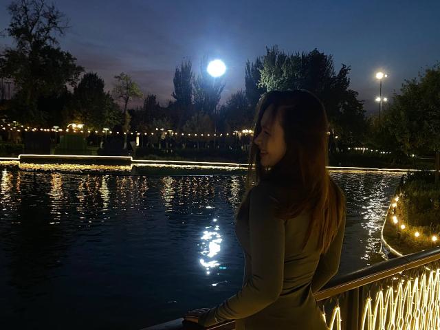 HotVibeK with brown hair, small tits, and a gray long-sleeve top, standing by a lit-up waterway at night, moon glowing above, reflection in water.
