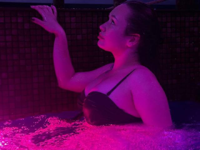 HotVibeK, with wet dark hair, is in a pink-lit hot tub, reaching up with her right hand, wearing a black strapless top, showing her boobs.