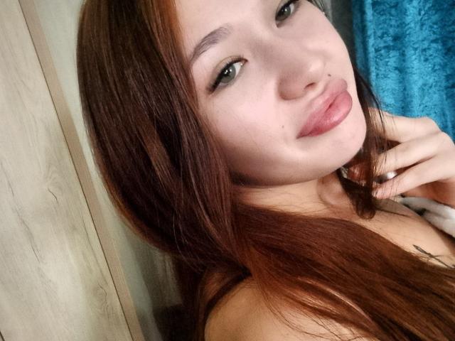 HotVibeK with long brown hair, pouty pink lips, and green eyes, giving a cute smile, touching her neck. She's got light skin and a subtle tattoo.