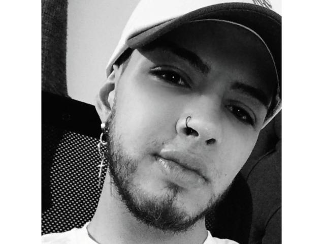 Sevven with a nose ring, beard, and cap, wearing a cross earring, gazing at the camera with a slightly parted lip, black and white selfie.