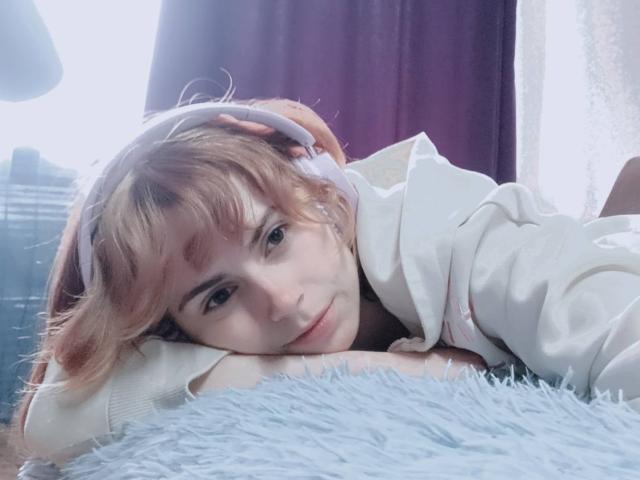 MireilleSpotwood with short, dirty blonde hair, white headphones, and a white hoodie, lying on a fluffy blue rug, face resting on her hands, smiling softly.