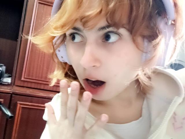 MireilleSpotwood with shocked expression, mouth open, hand raised, wearing pink headphones, light brown wavy hair, white shirt, wood cabinet in background, looking to the right, wide-eyed.
