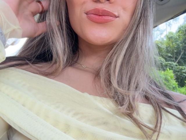 AdharaCooperr with her pouty lips, long grayish-brown hair, and yellow off-shoulder top, wearing a delicate necklace, her hand touching her head. Boobs partially covered.