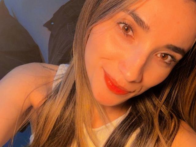 AdharaCooperr with long blonde hair, red lipstick, and a cute smile, showing her face and upper chest in a selfie, wearing a white tank top, looking at the camera.