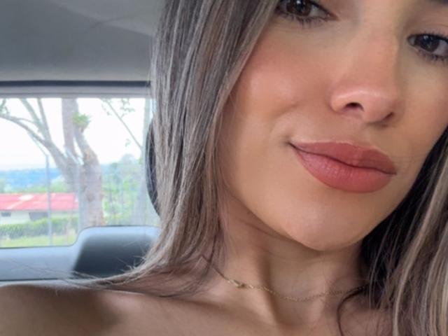 AdharaCooperr's face, showing light brown skin, long grayish-brown hair, and pinkish-brown lips, with a subtle smile, wearing a thin gold necklace, sitting in a car.