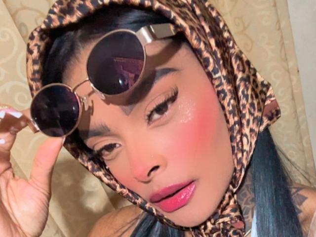 EmmaEbony with round sunglasses, pink lips, and a leopard print headscarf, holding glasses, has a pink blush on her face, and blue-black hair, looking slightly to the side.