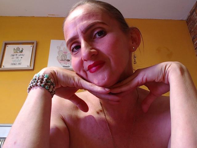 RubiAnderson with red lipstick, hands touching her neck, smiling, wearing a beaded bracelet, earrings, and a necklace, yellow wall, and framed artwork in the background.