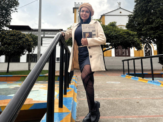 JessicaFiore with tan coat, black dress, patterned tights, and beanie, holding a book, standing on stairs, smiling, with black boots, in an outdoor setting, under a cloudy sky.