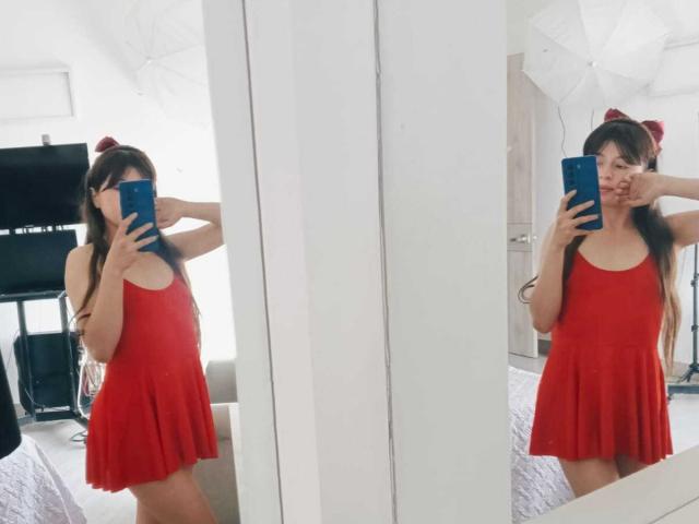 VioletaNova with long brown hair, wearing a red dress, taking mirror selfies, holding blue phone, covering mouth with hand, small ass, room with white walls and TV.