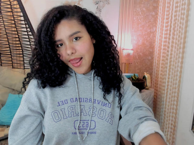 AriaVega with curly black hair, light brown skin, and small nose ring, wearing a gray "UCLA" hoodie, tongue slightly out, looking at camera. Soft light, cozy indoor setting.