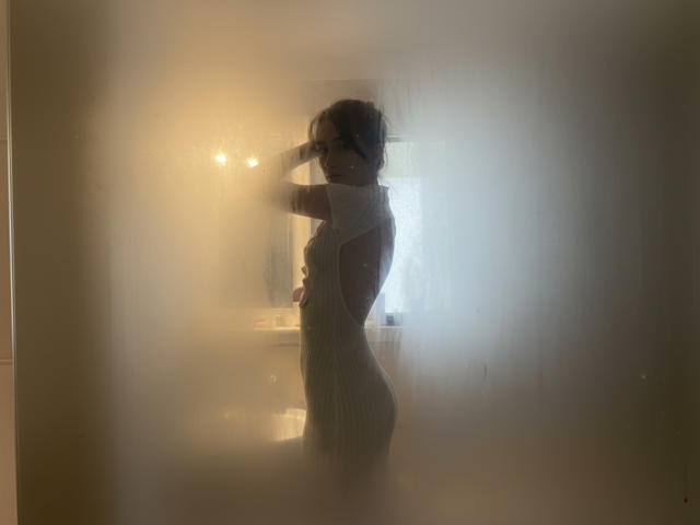 LuraSalminen, in silhouet, staand in een mistige kamer, gekleed in een strakke, witte jurk, armen omhoog, rug naar de camera, klein achterwerk, gemiddelde borsten, donker haar, een gloeiend licht achter haar..