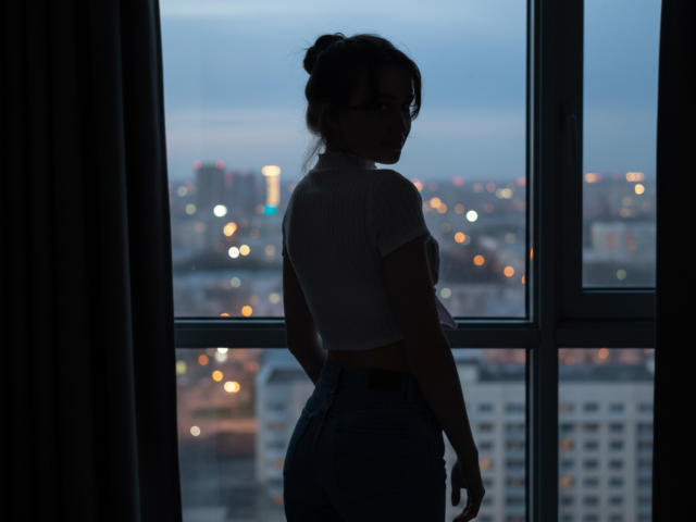 Silhouetted LuraSalminen stands in front of a window, back to the camera, wearing a white crop top and blue high-waisted jeans, city lights glowing in the evening.