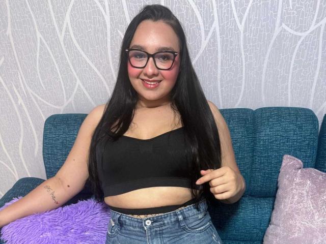 ValeryAndreas with long black hair, glasses, pink blush, black sports bra, showing her tits, and blue jeans, sitting on a blue couch with purple and pink pillows.
