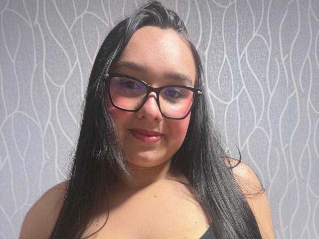 ValeryAndreas has black hair, glasses, pink blush on cheeks, and red lipstick, with medium-sized tits and a cute smile. She's looking at the camera, smiling softly.