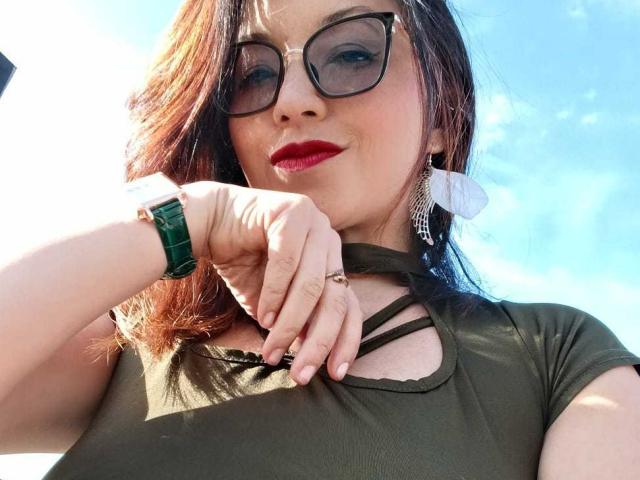 AngelinaBlanche has red lipstick, green glasses, and a green watch. She's wearing a green top with a cutout, and her hand is near her chin, showing a ring.