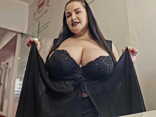 GeorgiaLerox with big tits in a black lace bra, holding up a sheer black top, looking down with a slight smile, long black hair, dark lipstick, and red nails.