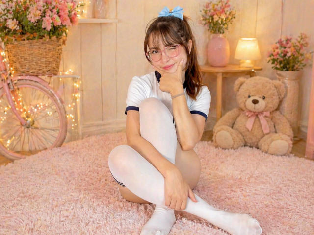IsabelaP with glasses, white t-shirt, and white thigh-high socks, sitting on pink shag rug, smiling, hand on chin, brown pigtails with blue bow, pink teddy on right.