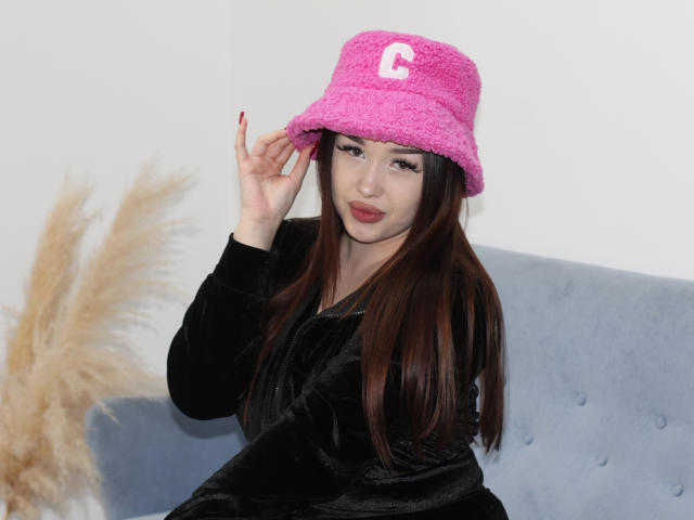 LexiaEllon with long brown hair, wearing a pink fuzzy hat with a white letter 'C', black velvet robe, red nails, sitting on a gray couch, touching hat.