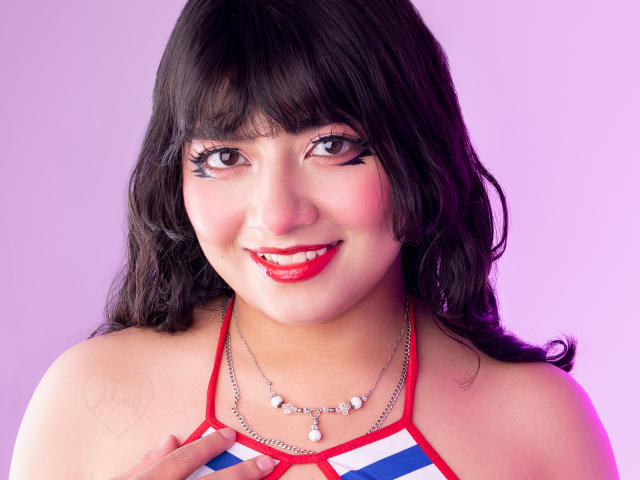 NekoPam has curly black hair, red lipstick, and a smile. She's wearing a striped top, red halter, and a necklace with pearls. She's touching her chest.