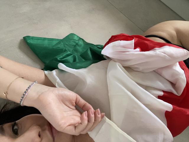 Dionisia laying on floor with Italian flag, white shirt, red and green, black thong, visible ass, bracelet on left wrist, holding shirt, partial face, brunette, relaxed.