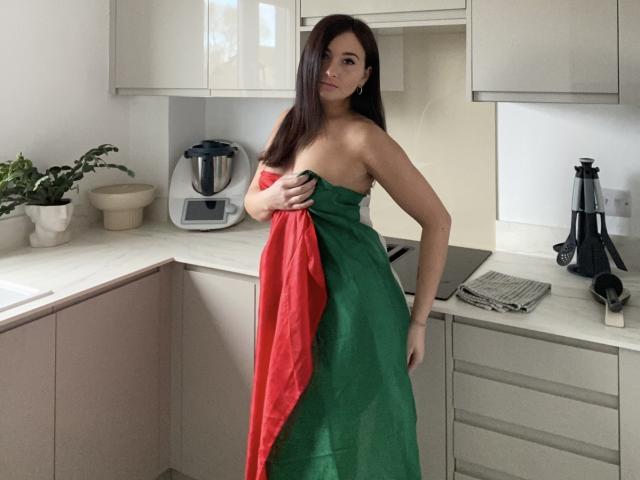 Dionisia with long brown hair stands in a kitchen, holding a green and red cloth over her bare tits, wearing it like a dress. She's looking at the camera.