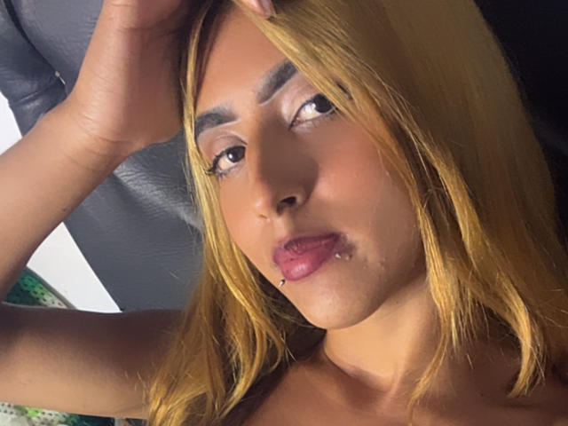 SophiaForeest hat eine hellbraune Haut, blondes Haar und eine Lippenpiercing. Sie schaut zur Seite und hat ihre Hand im Haar, wobei ein kleiner Schönheitsfleck sichtbar ist..