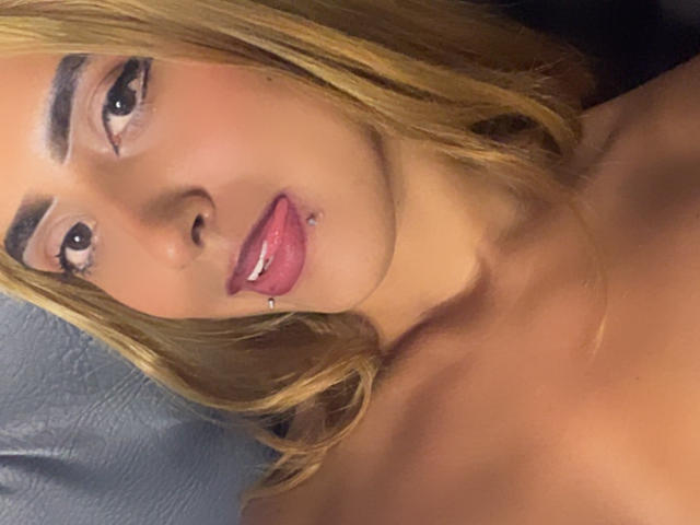 SophiaForeest a les cheveux blonds, des sourcils foncés, des lèvres roses et brillantes avec un piercing, et une légère peau bronzée. Ses yeux sont dirigés vers l'appareil photo. Ses épaules sont visibles..