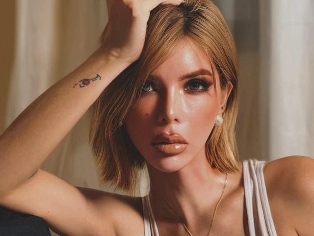 HotCumAdriana69x has blonde hair, a tattoo on her right arm, and is wearing a white top with a gold necklace. She has full lips and is looking at the camera.