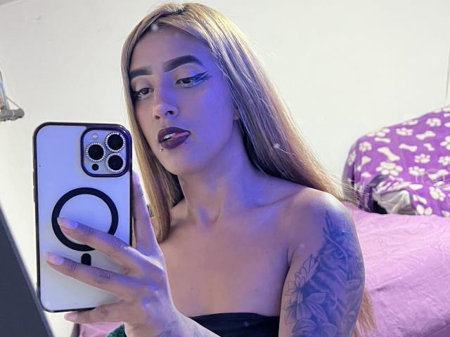 SophiaForeest mit langen blonden Haaren, schwarzem Lippenstift und einem Tattoo auf ihrem rechten Arm, macht ein Selfie mit ihrem iPhone vor einem Spiegel, wobei ihre Zunge leicht herausragt..
