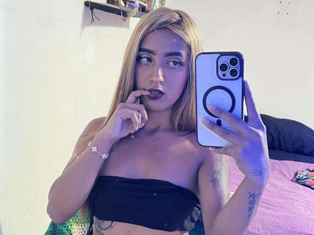 SophiaForeest, com cabelo loiro, lábios carnudos, dedo na boca, segurando um telefone, top preto sem alças, tatuagens no braço, tirando uma selfie no espelho, luz azul no lado direito..