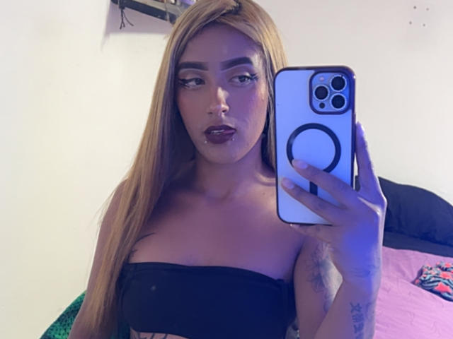 SophiaForeest, avec de longs cheveux blonds, portant un haut noir sans bretelles, tenant un iPhone avec une coque décorée, regardant la caméra, montrant des tatouages sur son bras et son poignet..
