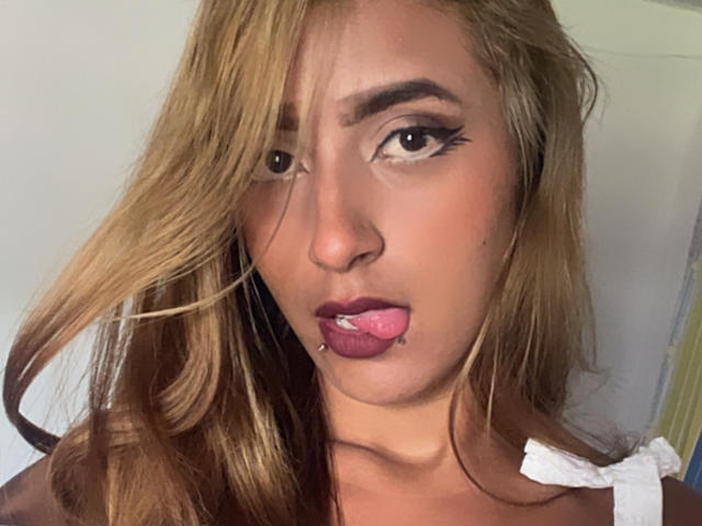 SophiaForeest hat blondes Haar, dunklen Eyeliner und steckt ihre Zunge heraus, wobei sie ein Lippenpiercing trägt. Sie trägt ein weißes Oberteil und schaut direkt in die Kamera..